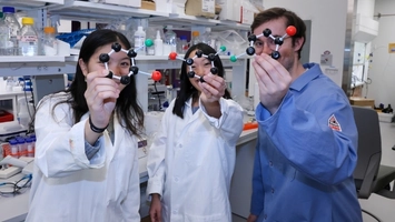 Three researchers in lab coats holding molecular model structures in a well-equipped laboratory setting